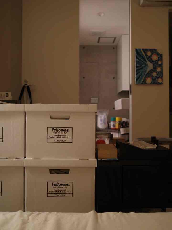 白を基調としたコンパクトな寝室、ベッドの前にBANKERS BOXの収納ボックスが積まれ、ナイトテーブルの上にティッシュケースが置かれた落ち着いた空間