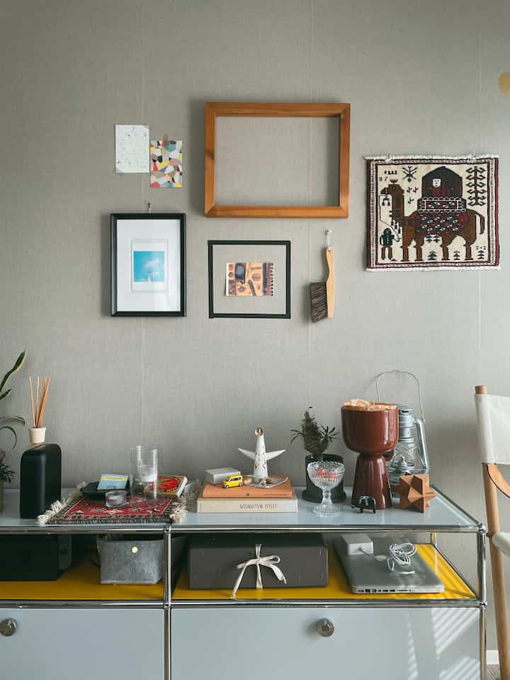 ベージュトーンの壁に多様な額縁が飾られたナチュラルスタイルの空間、食卓の上には様々な小物と本が置かれ、温かみのある雰囲気のあるインテリア