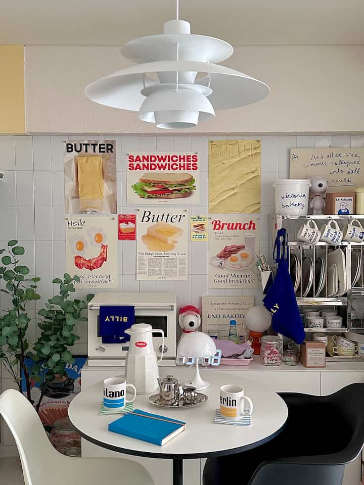 White-toned kitchen and dining room featuring a round dining table and pendant light centrally, with various posters arranged on the wall creating a cozy space