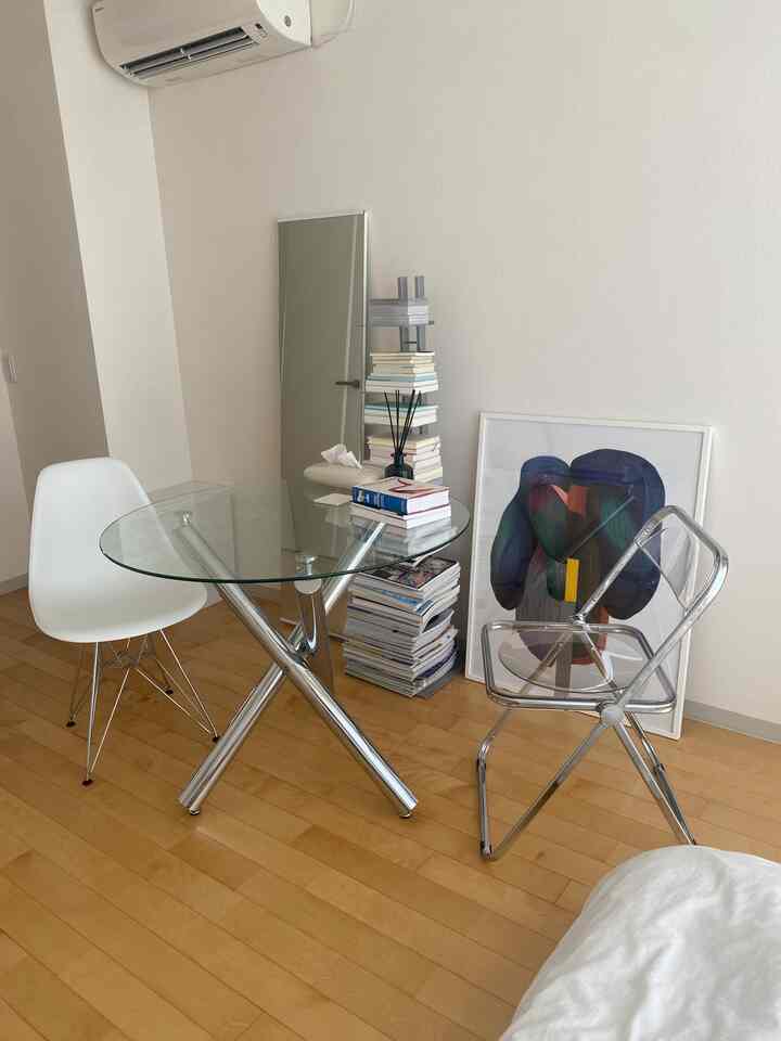 明るい木目調の床と白い壁の小さな空間に、透明な椅子と円形ガラステーブル、抽象的なポスターが配置されたミニマルなリビング