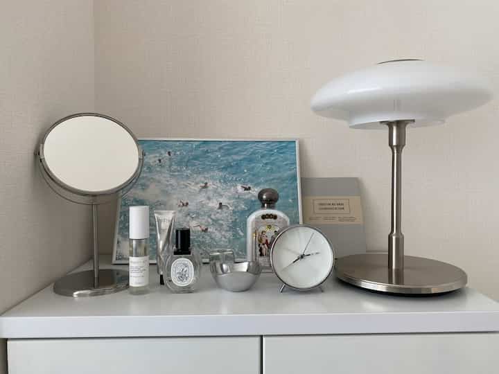 White and silver toned nightstand featuring a round mirror, poster, clock, and table lamp in a clean setting