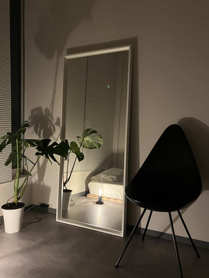 Minimal bedroom with white walls and floor, large mirror and black armchair, accompanied by green potted plant