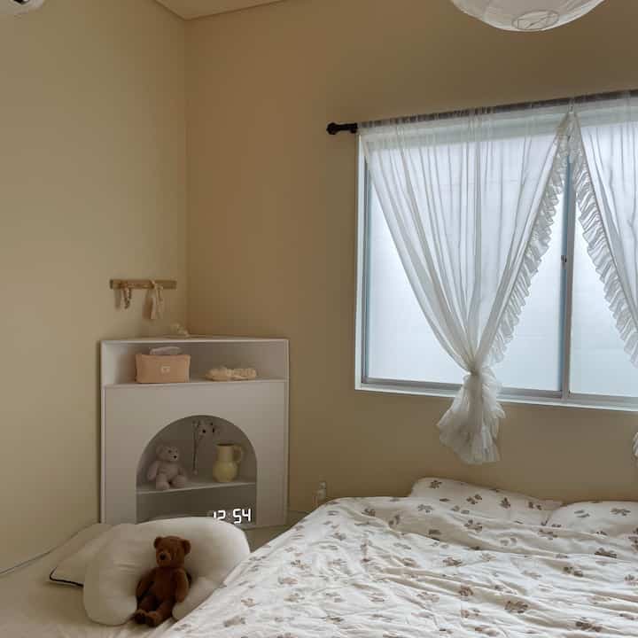 ベージュとホワイトの色調の寝室、くまのぬいぐるみとフリル付きのカーテンが印象的な空間