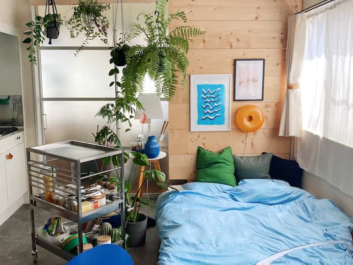Bright natural-tone studio apartment featuring blue bedding bedroom, stainless kitchen cart, and abundant plants creating cozy ambiance