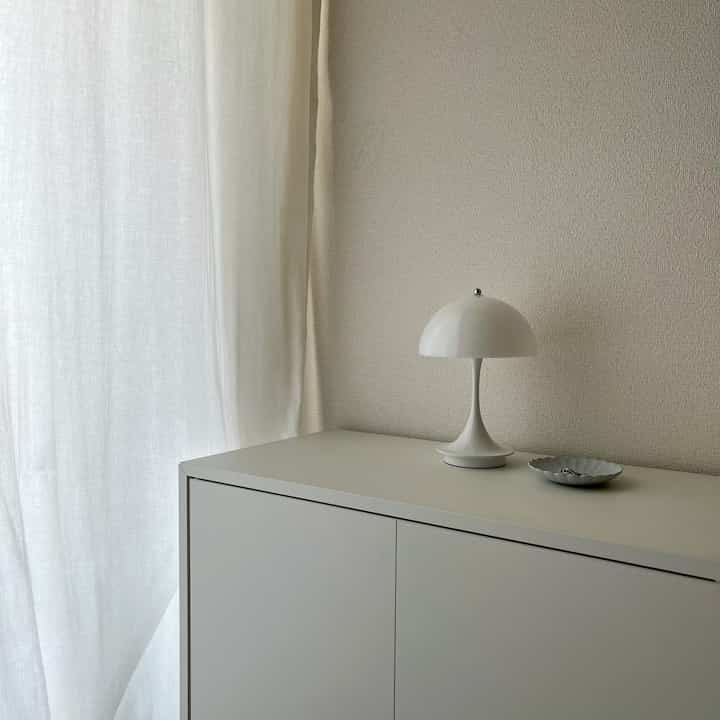 白とナチュラルな色調のワンルーム、シンプルなキャビネットの上にテーブルランプと皿が置かれた落ち着いたインテリア