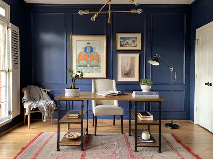 Navy-colored wall home office featuring a wood tone desk and cream chair with a calm and sophisticated atmosphere
