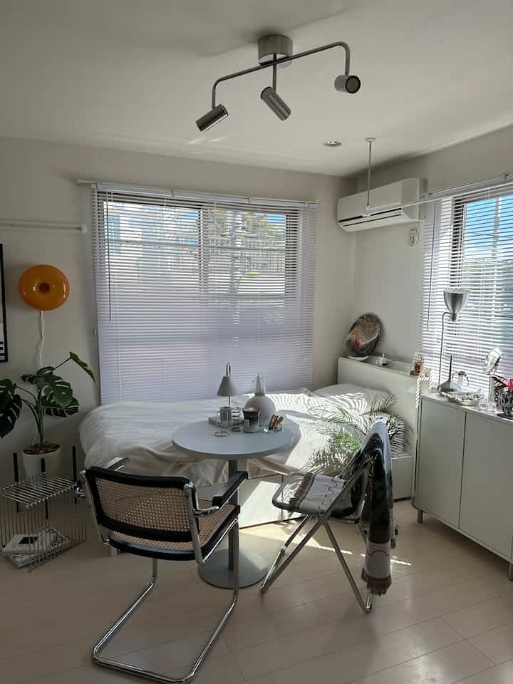 White-toned studio bedroom featuring white furniture, blinds, and mixed wood-metal chairs in a clean space