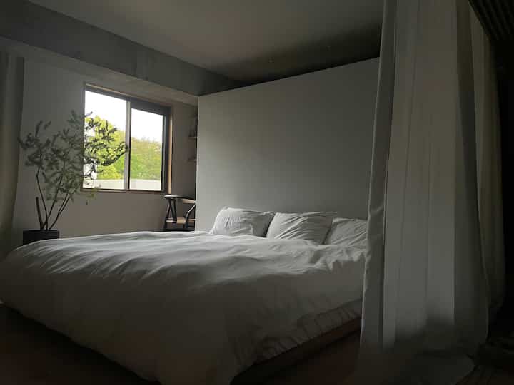 Calm toned bedroom featuring a large bed with white bedding centered and a black chair by the window providing natural light