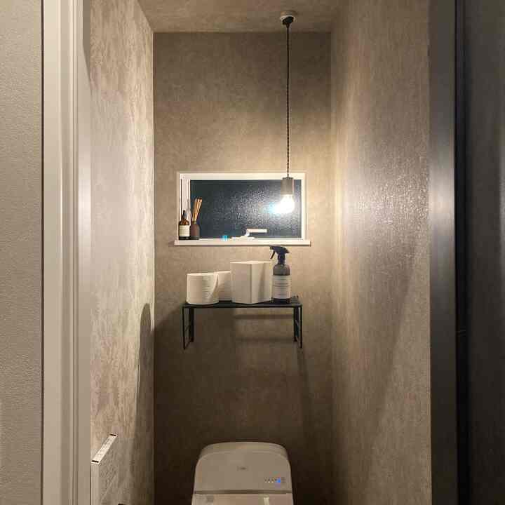 ベージュ色の壁と黒のペンダントライトがあるシンプルなトイレ、オープン棚に収納アイテムが配置された雰囲気のあるインテリア