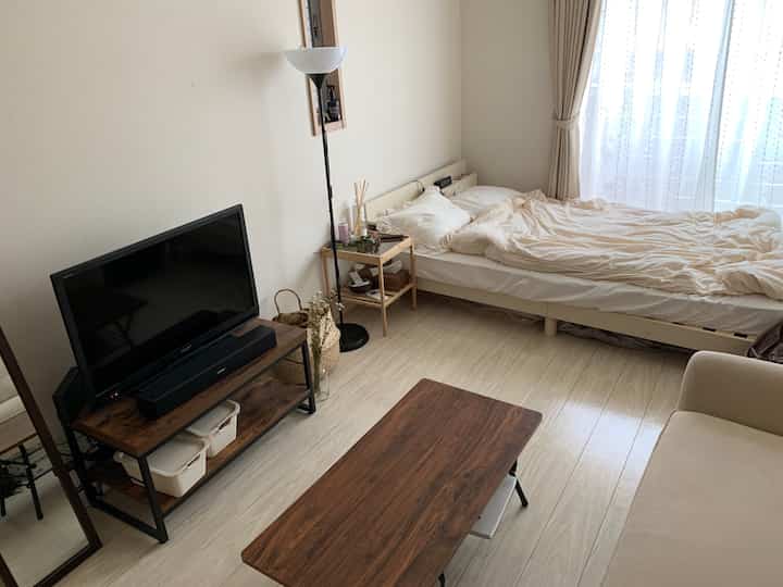 ホワイトとライトブラウンのトーンで統一されたワンルームのリビング兼寝室、テレビ台とベッド、ソファが配置された落ち着いたナチュラルな空間