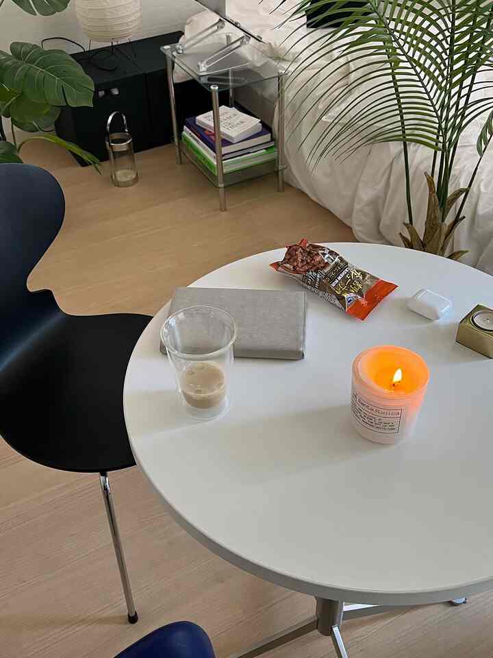 自然光が差し込むウッドトーンの床が印象的なシンプルな小さな部屋、白い丸テーブル上にキャンドルとガラスのカップ、黒い椅子と植物が配置された雰囲気のあるインテリア