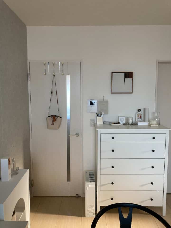 明るいホワイトトーンの玄関、ドアにベージュ色のドアフックとバッグが掛けられ、右側にホワイトのチェストが配置されたシンプルなインテリア