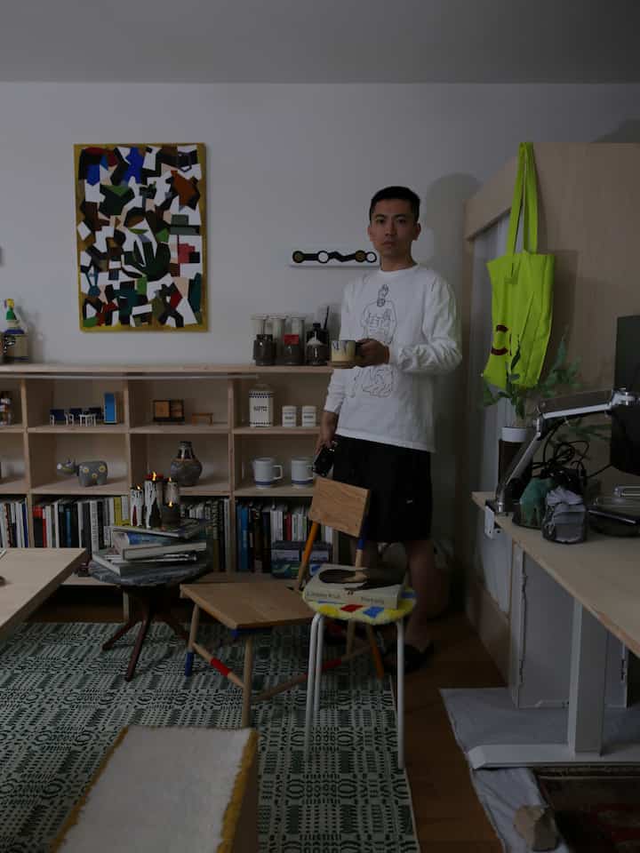 緑のパターンのラグが敷かれたワンルームのリビング、壁のアート、棚、机、マグカップを持つ男性がいるモダンなインテリア