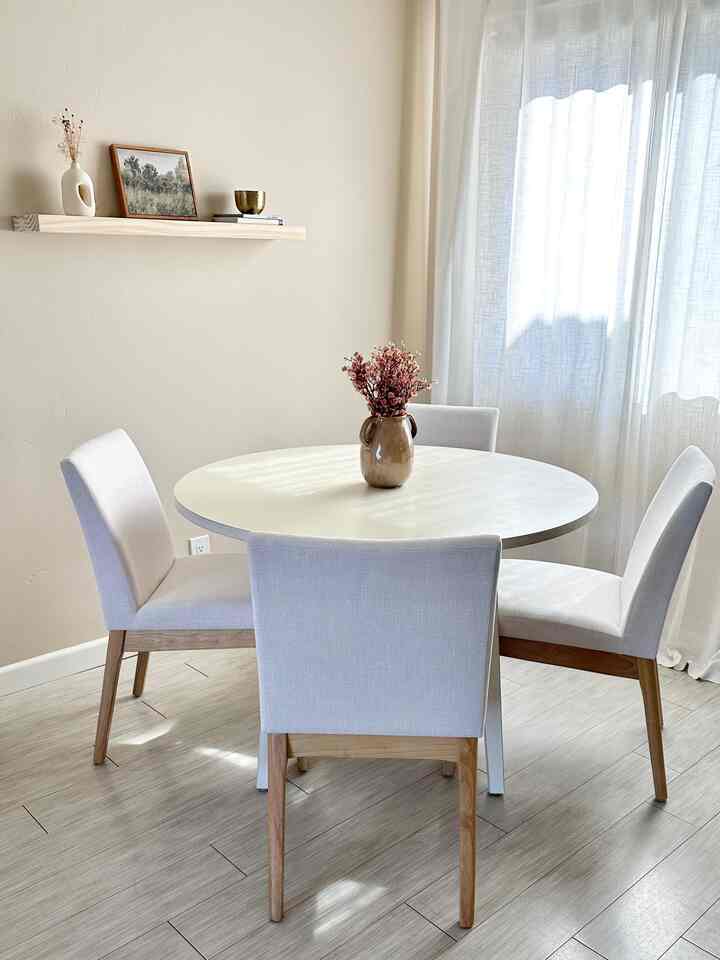 Bright natural-toned dining room featuring a white round table with four chairs and a floating wooden shelf with wall decor