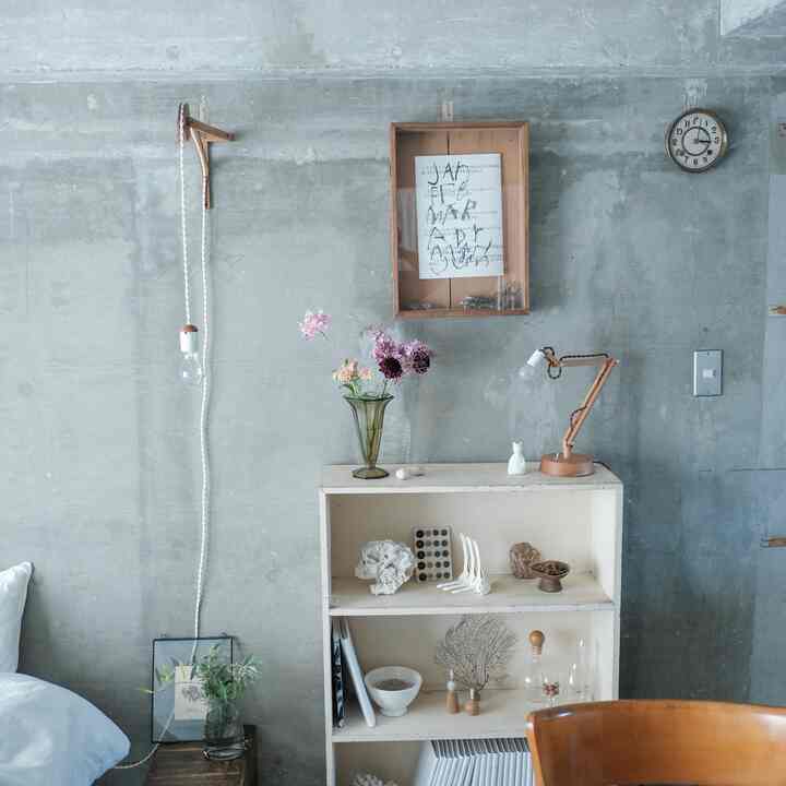 ナチュラルなベージュトーンとコンクリート壁が調和するワンルームの内部、書棚とテーブルランプが配置されたシンプルな空間