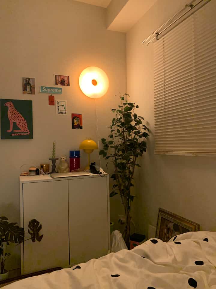 Warm yellow lighting illuminates a white-toned studio apartment bedroom featuring plants and a simple storage cabinet, creating a cozy atmosphere
