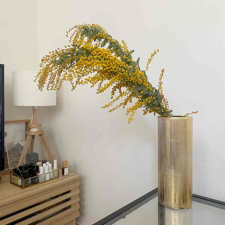 白い壁と木目調のチェストの上に、白いテーブルランプと金色の花瓶が置かれ、黄色い花が飾られた雰囲気のあるインテリア