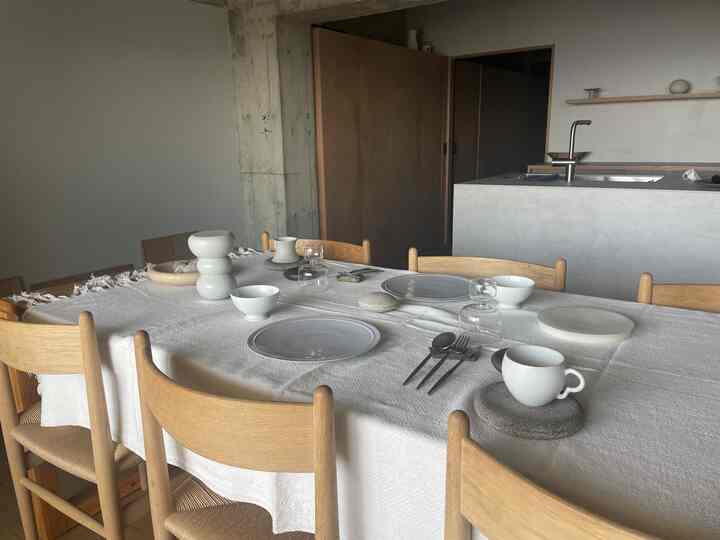 白いテーブルクロスと木目調のダイニングチェアが配置されたシンプルなダイニング、背景にモダンなキッチンが見える