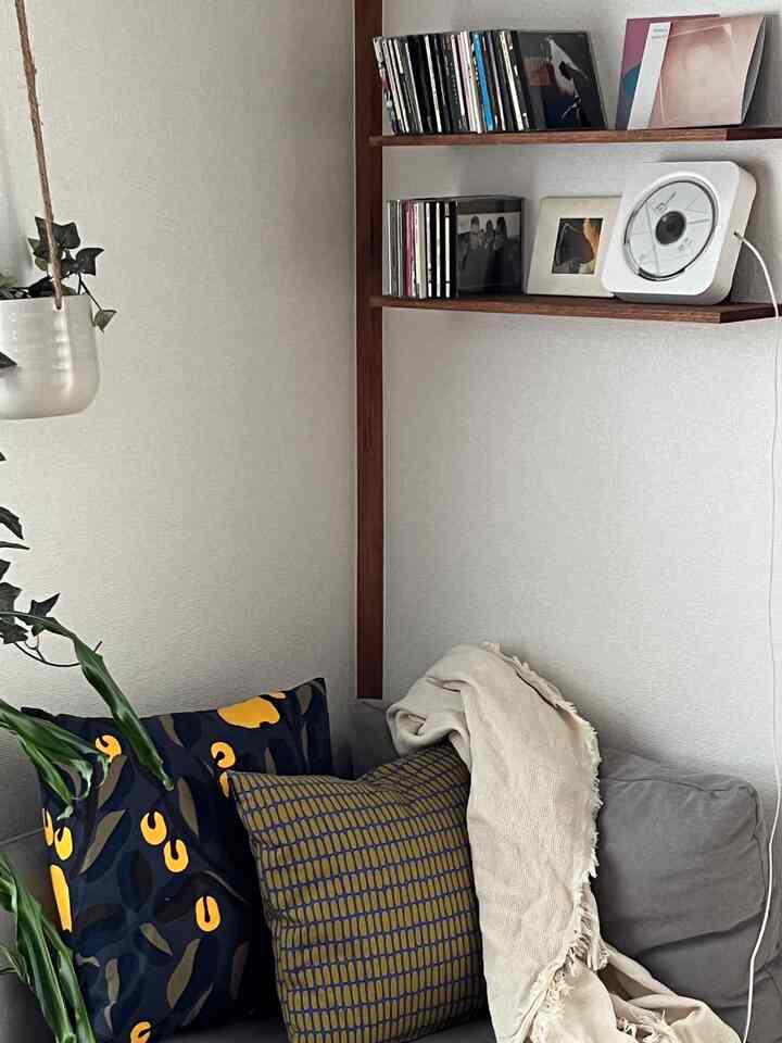 白い壁と木目調の棚がある一人暮らしのリビング、グレーのソファとクッション、植物が配置されたシンプルで自然な雰囲気のインテリア