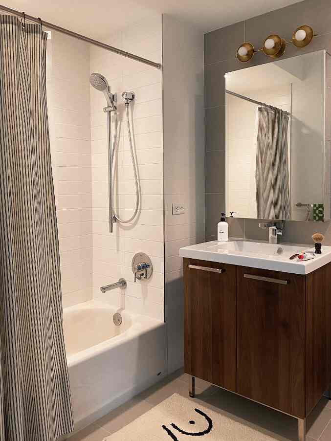 White and wood-toned bathroom featuring a cream bath mat, striped shower curtain, and wooden vanity with mirror, creating a clean and warm atmosphere