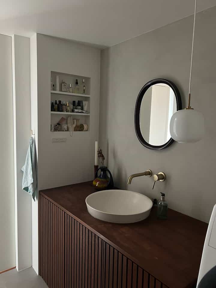 グレーの壁とダークブラウンの木製洗面台があるナチュラルテイストのバスルーム、陶器製洗面器と楕円形の鏡、ペンダントライトが調和する空間