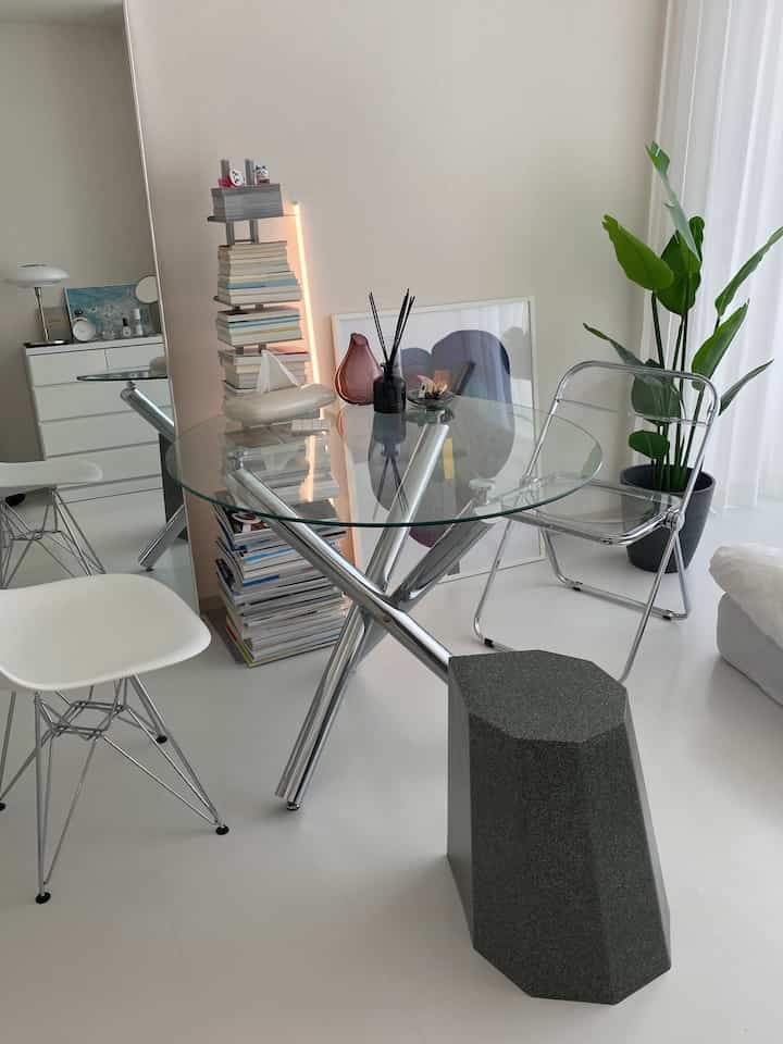 白い床とモダンな家具があるカフェ風リビング、透明なテーブル、縦に積まれた本棚、緑の植物が配置された雰囲気のある空間