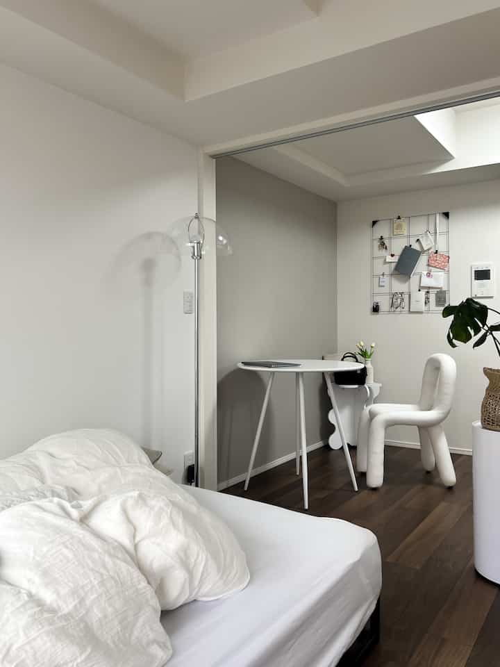 A modern, clean bedroom with dominant white and natural brown tones featuring a small round desk and chair