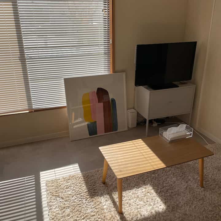 ナチュラルカラーのリビング、白いキャビネットの上にテレビがあり、木目調のコーヒーテーブルとブラインドが配置された落ち着いた空間