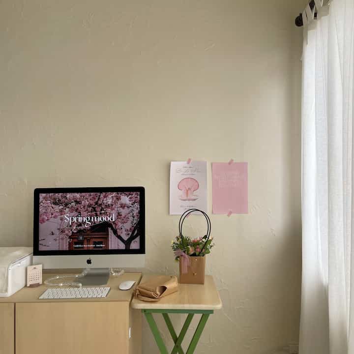 明るいベージュの壁と木製の机があるコンパクトなワークスペース、壁にはピンクのポスターと植物が配置されたインテリア