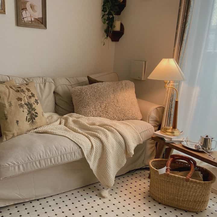 Warm beige-toned living room featuring a two-seater sofa, a classic table lamp, and a knitted blanket creating cozy ambiance
