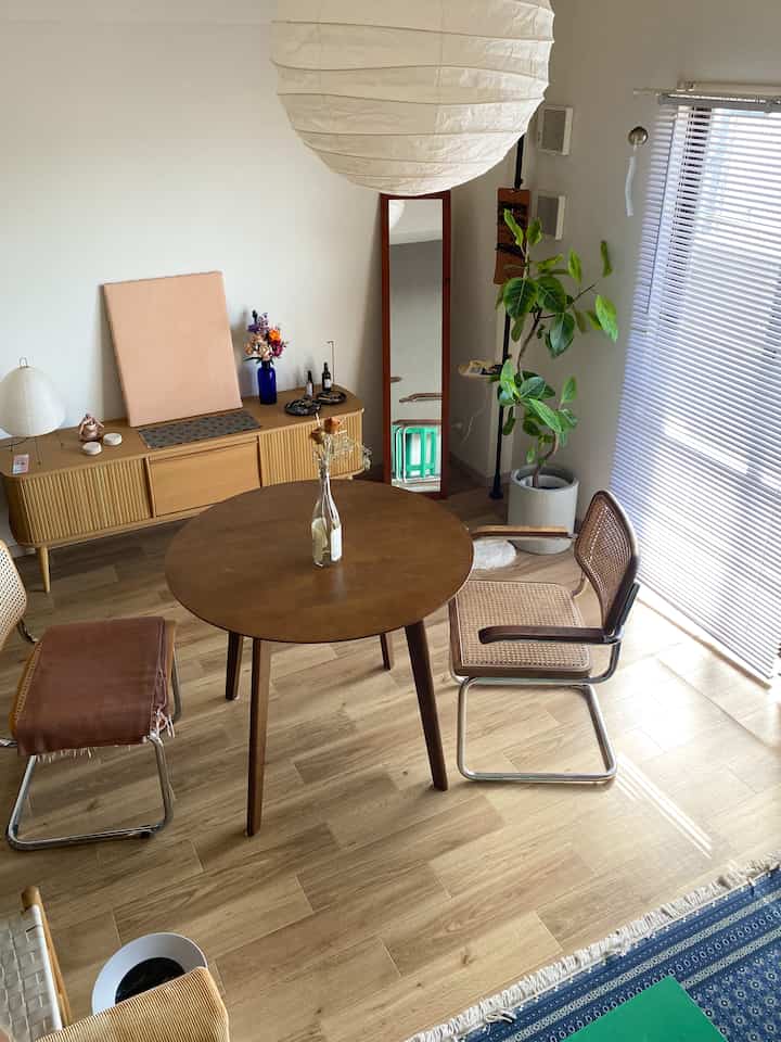 ナチュラルトーンのリビング、丸い木製ダイニングテーブルとアームチェア、大きなペンダントライトとブラインドが調和した洗練された空間