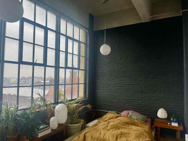 Bedroom with black brick accent wall and yellow bedspread, featuring multiple plants by large grid windows creating a cozy atmosphere