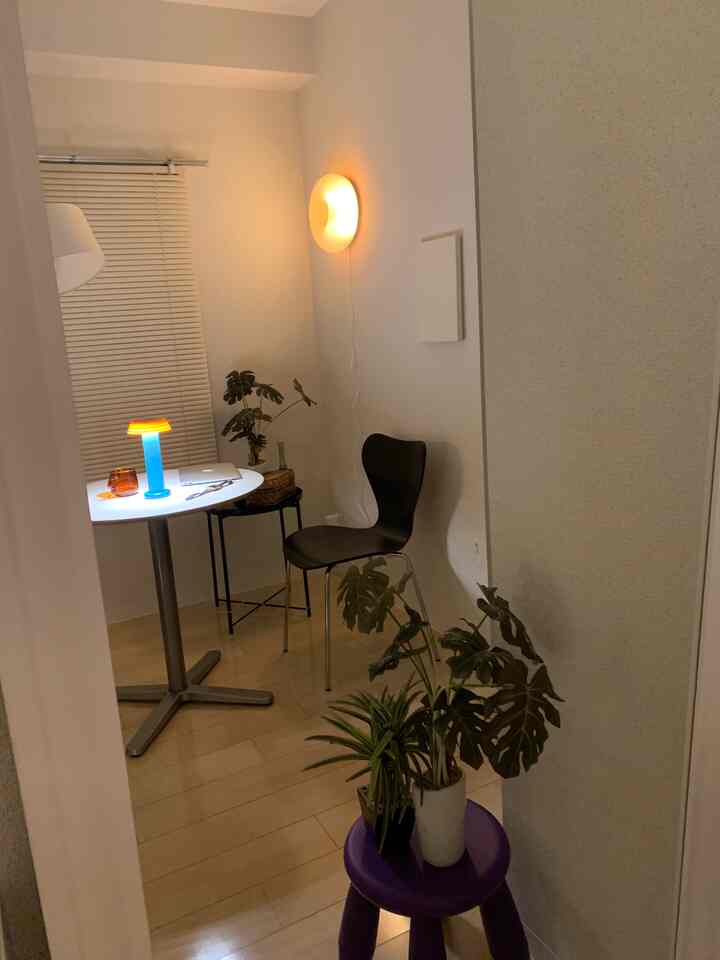 Light gray-walled small studio dining area featuring a round dining table, black mid-century modern dining chair, vibrant table lamp, and indoor plants creating cozy atmosphere