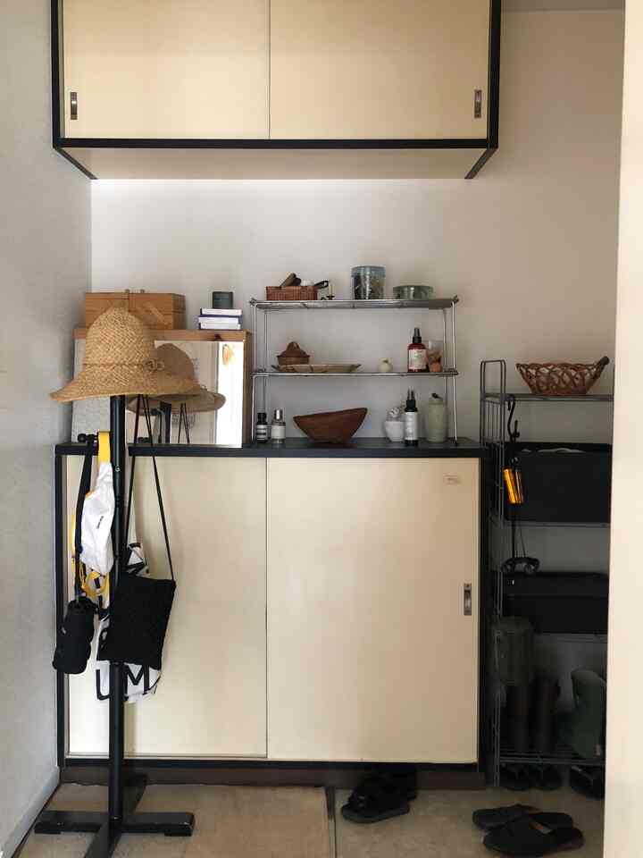 ベージュ色調のコンパクトな玄関、黒い棚と帽子スタンド、収納家具が配置されたやや暗い空間