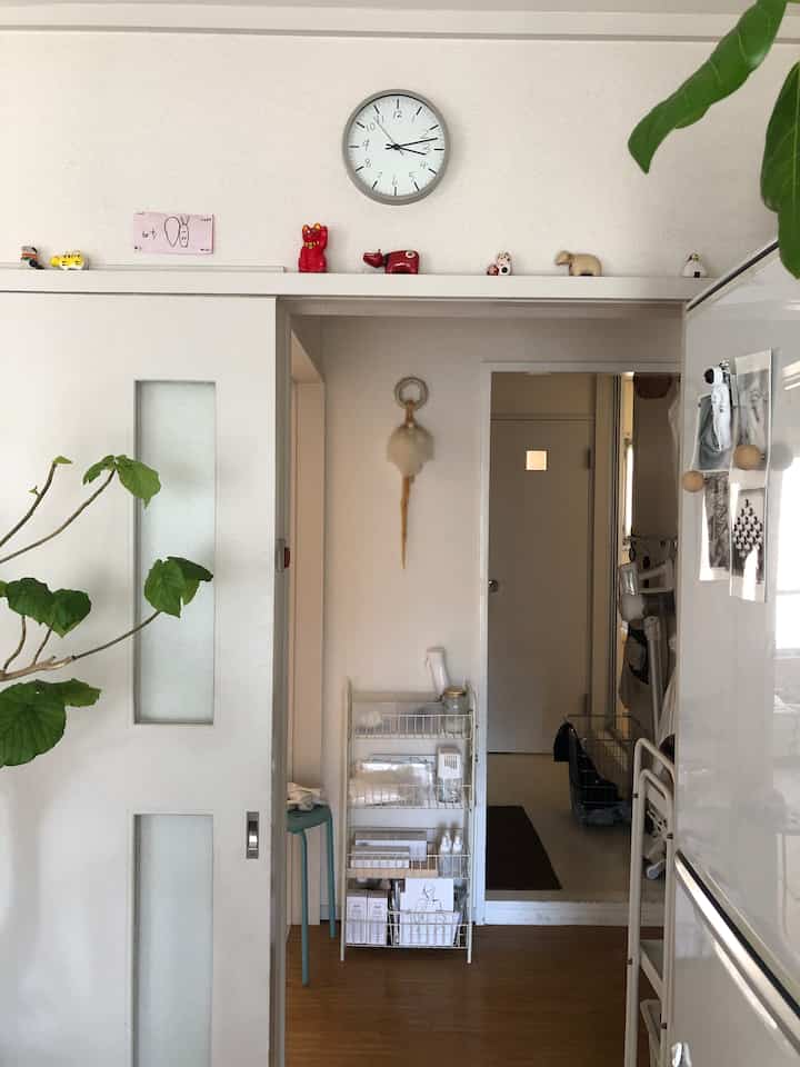 白い壁と木の床のリビング入口、緑の植物とスツールが配置された落ち着いたミニマルな雰囲気の空間