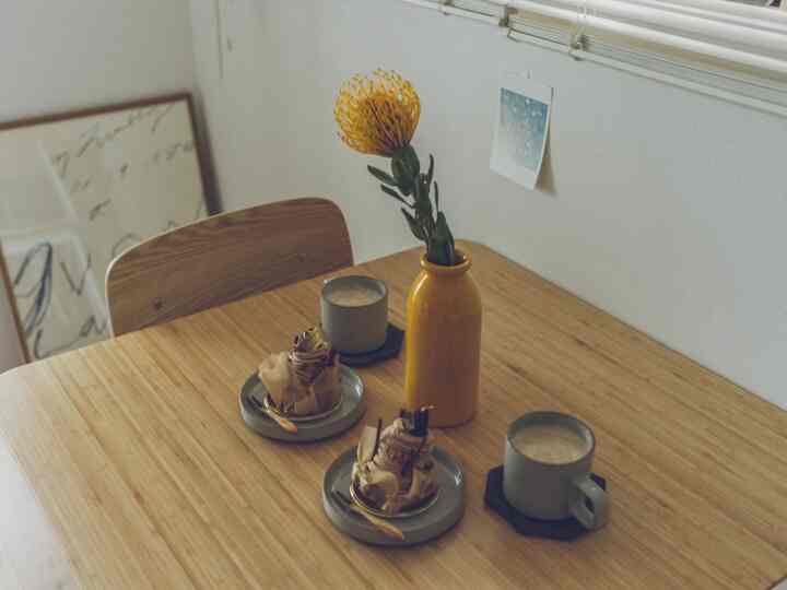 ナチュラルな色調のダイニング、中央の木製テーブルに黄色い花瓶とグレーの食器が配置された雰囲気のあるおうちカフェ空間