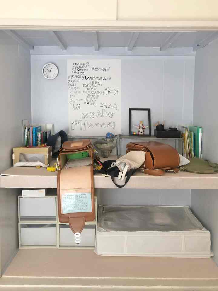 白と茶色のトーンの子供部屋の棚、中央に二つのランドセルと本、収納ボックスが散らかった印象のあるインテリア