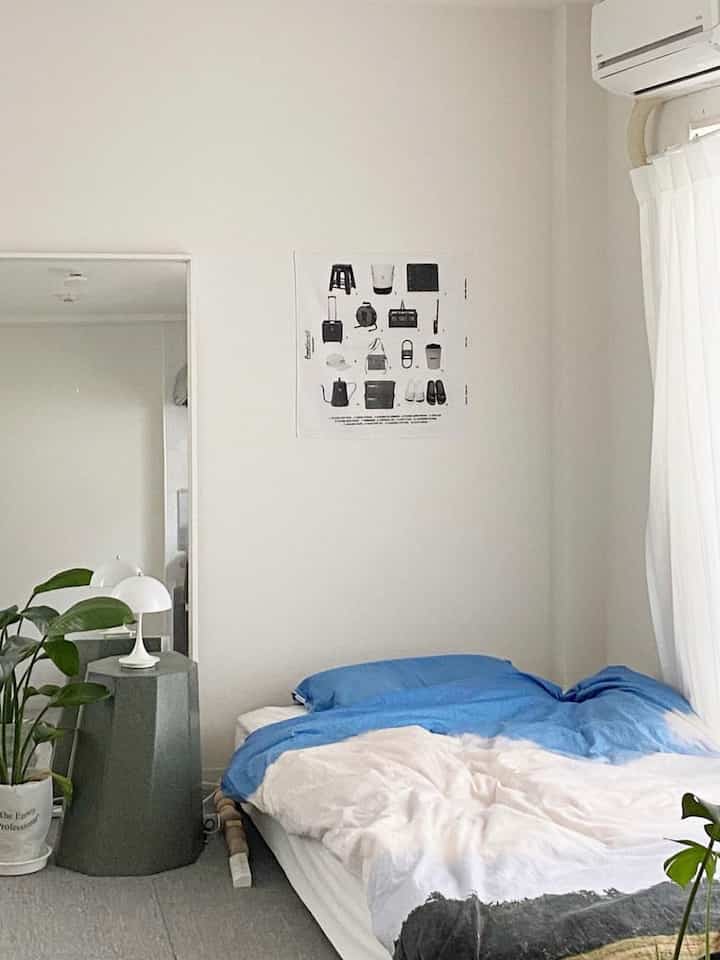 白と青の色調の寝室、木製ベッドとスツール、ポスター、植物が配置されたシンプルで落ち着いたインテリア