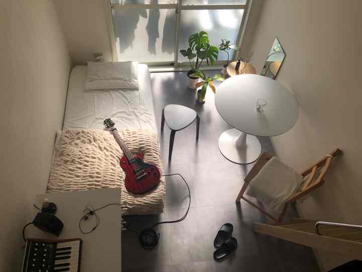 白とダークグレーのトーンによる小さなワンルーム、ベッド、ダイニングテーブル、スツール、植物が配置されたミニマルで自然なインテリア