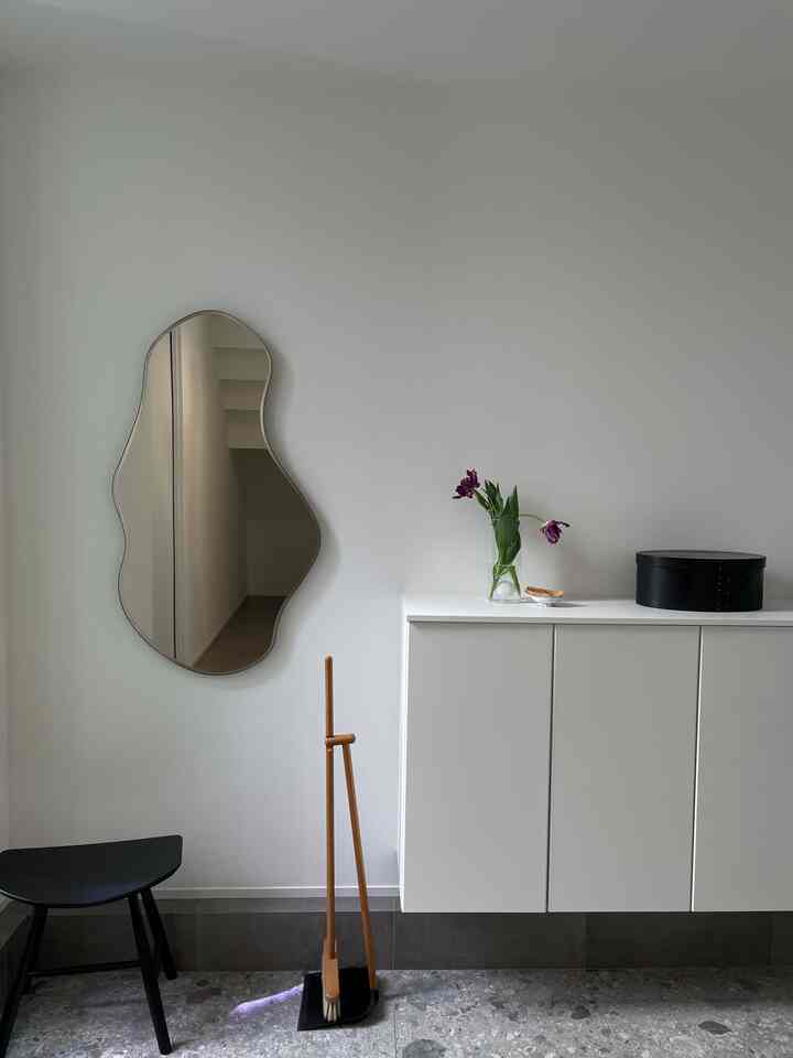 白い壁とキャビネットがあるモダンでシンプルな玄関、鏡と黒いスツール、植物が配置された雰囲気のあるインテリア