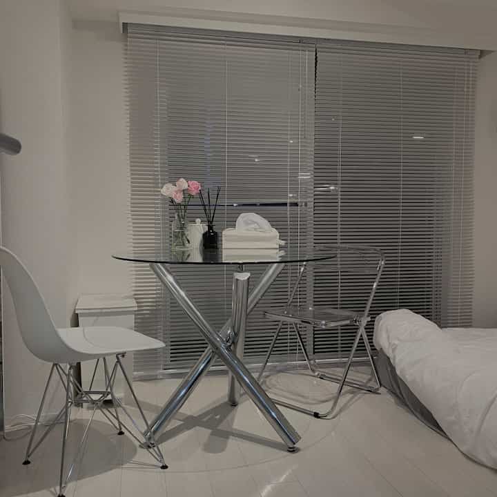 White and gray toned small bedroom space featuring aluminum blinds and transparent dining chairs in a simple interior