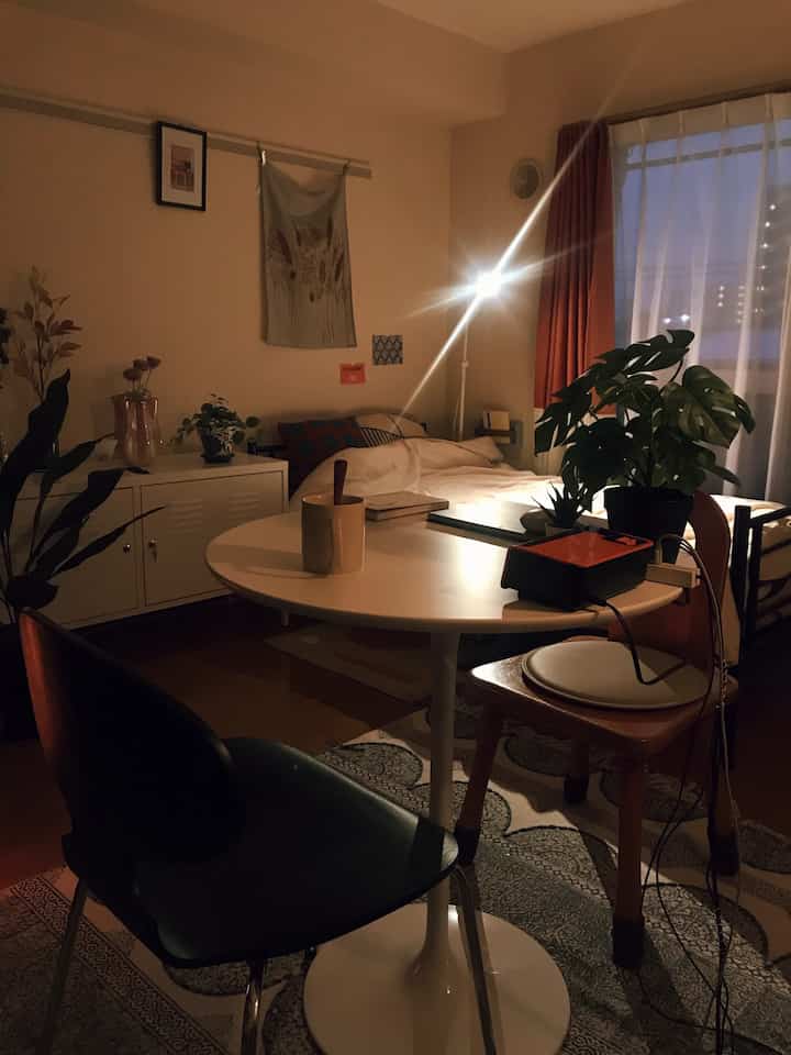 暖かいホワイトとブラウンのトーンの一人暮らし用寝室、中央に白い円形ダイニングテーブルと植物が配置された居心地の良い空間