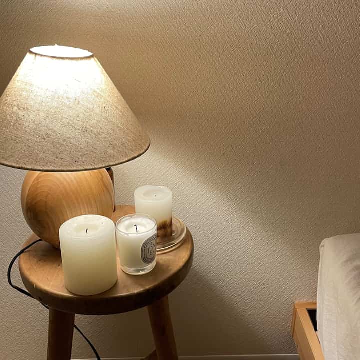アイボリー色の壁と木目調のサイドテーブル、キャンドルとテーブルランプが配置された、落ち着いた寝室の一角。