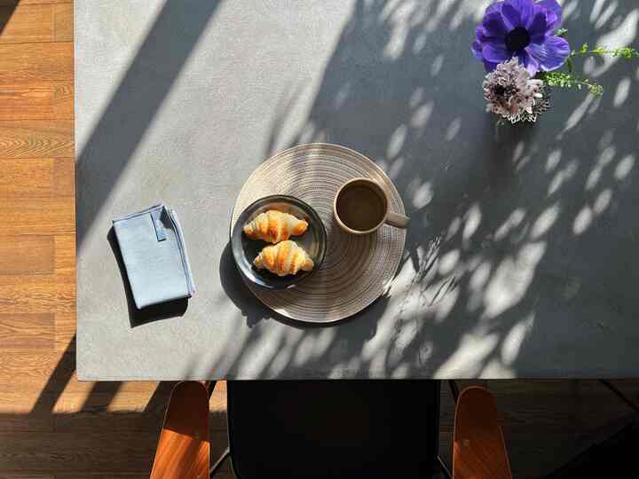 ウッドトーンの床とモダンなグレーのダイニングテーブルにおうちカフェのセッティングがあり、クロワッサンとコーヒー、花瓶が配置された雰囲気のあるインテリア