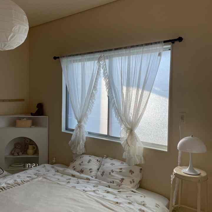 ベージュの壁と白いシアーカーテンがある小さな寝室、中央にベッドとナイトテーブル、テーブルランプが配置された落ち着いた空間