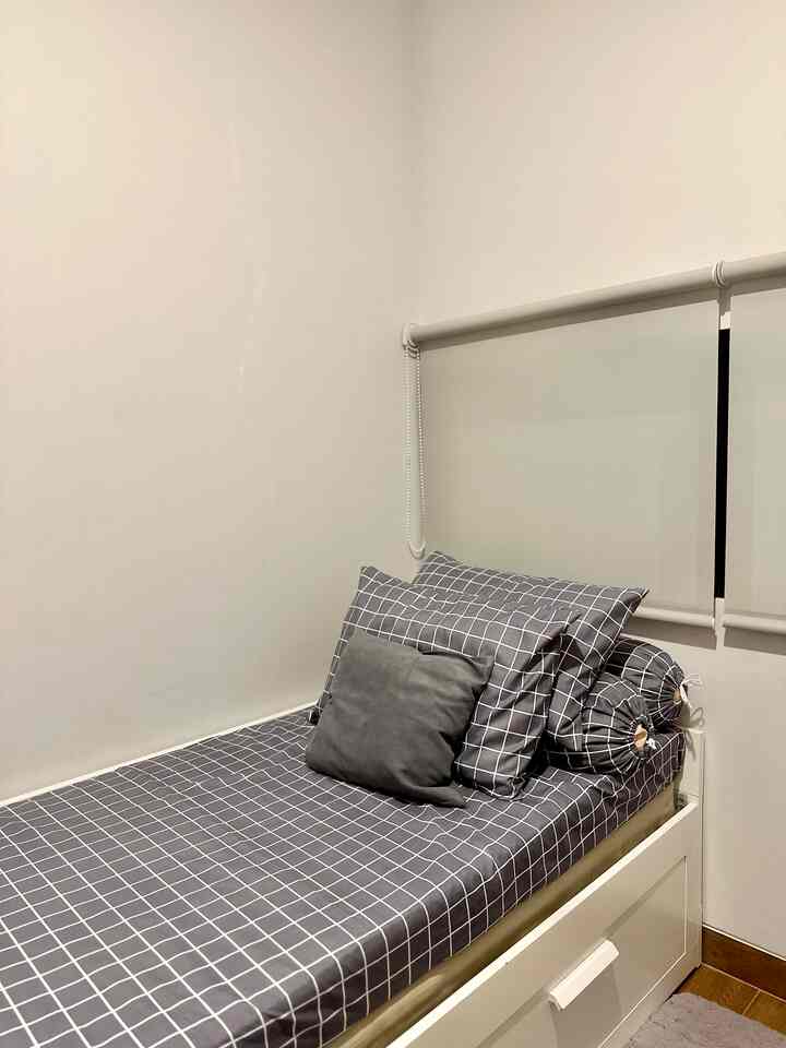 White and gray toned kids' bedroom featuring a simple bed with grid-pattern bedding and roller blinds in a compact space