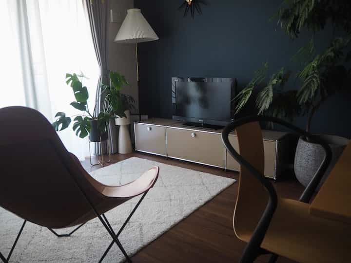 Natural color toned living room with dark blue accent wall, featuring TV stand, lounge chair, floor lamp, and plants in a modern setting