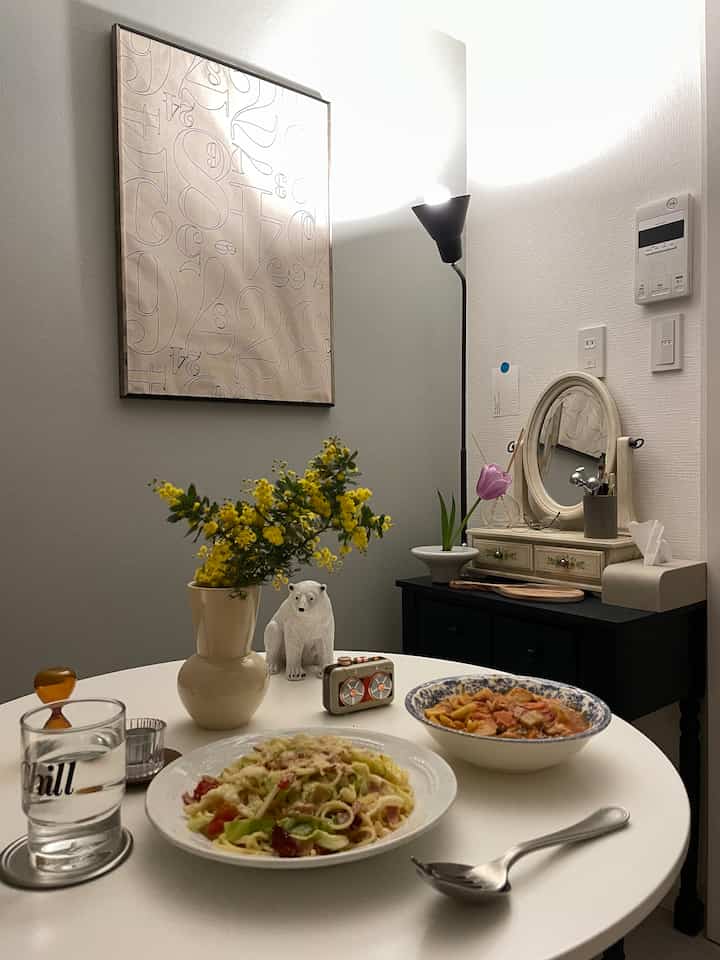 白とベージュのトーンが広がる狭い部屋に、丸テーブルとフロアランプ、黄色いミモザの花が置かれた雰囲気のあるインテリア