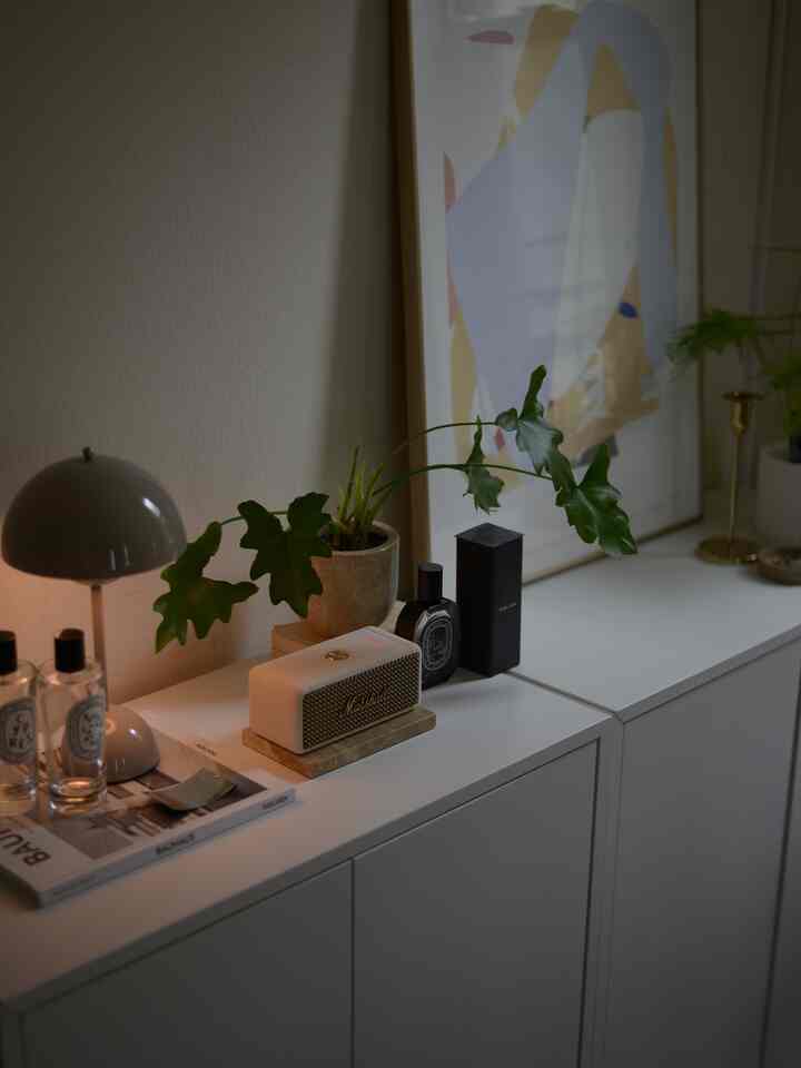 ホワイトトーンのキャビネット上にモダンなテーブルランプ、大きな室内植物、マシャルのブルートゥーススピーカーが配置されたミニマルな空間