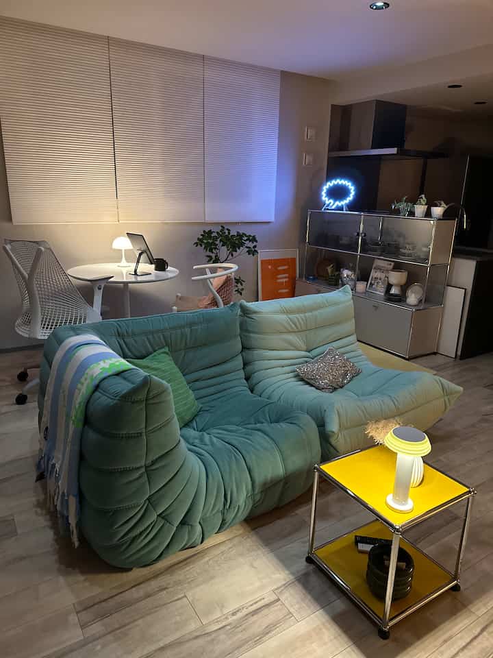 白と緑のトーンのリビング、中央にティールのセクショナルソファと黄色のサイドテーブルが配置された落ち着いた間接照明のある雰囲気のあるインテリア